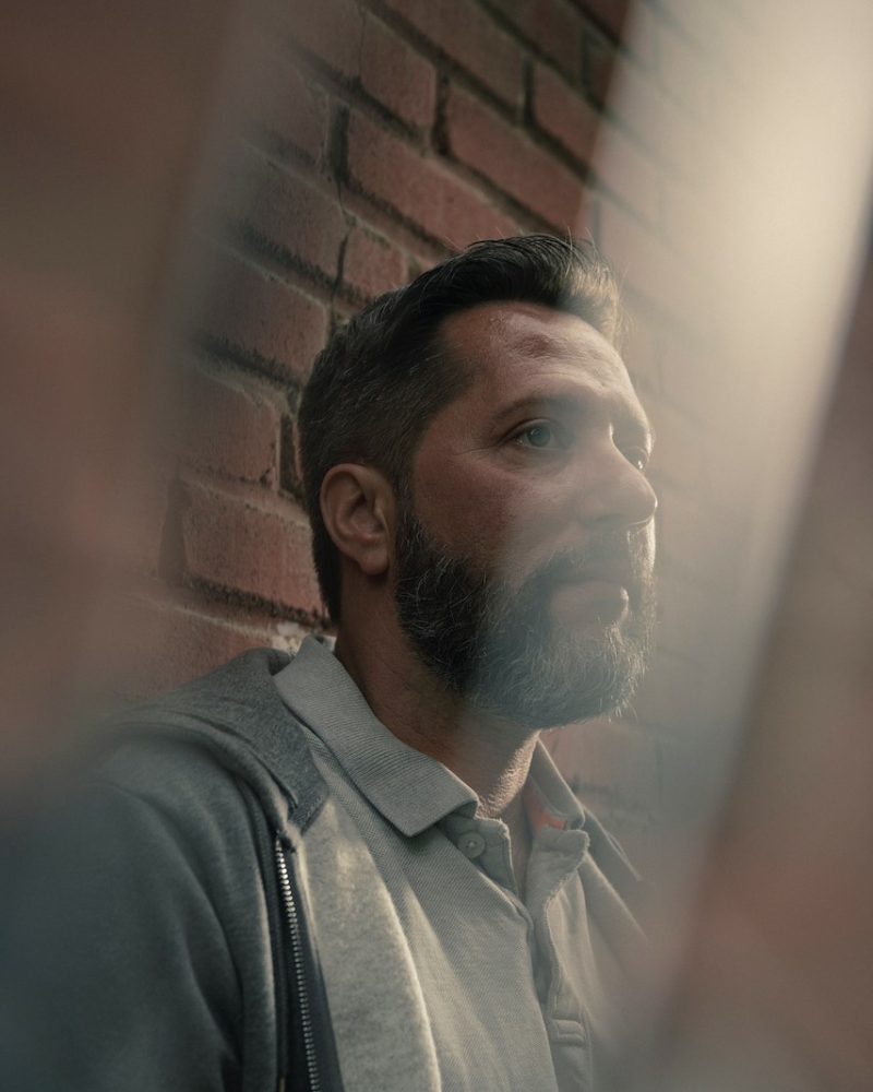 Homme à barbe, chemise grise, hoodie gris, contre mur de briques rouges